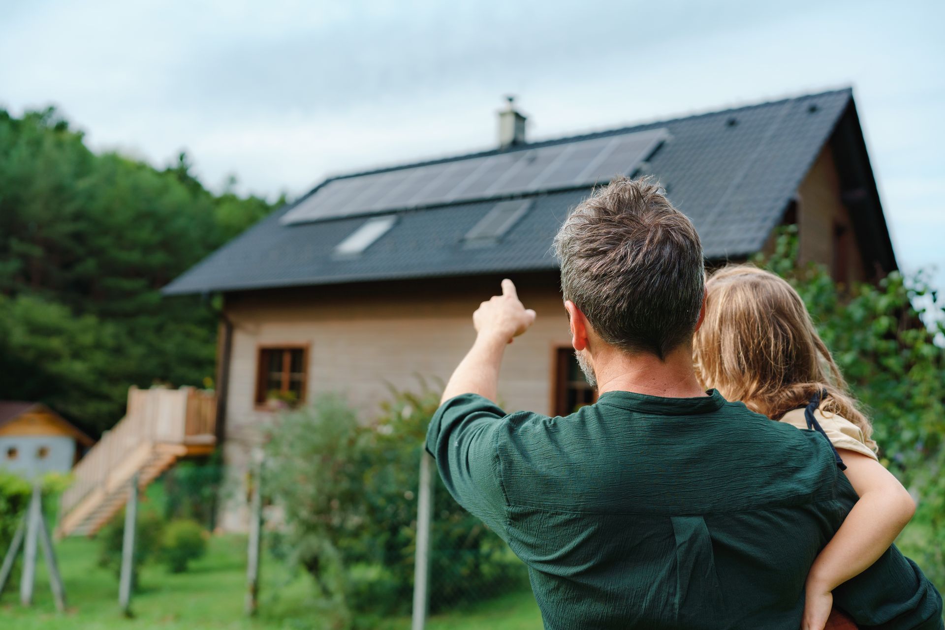 A person holding a child on their shoulder points toward a house with solar panels on its roof in a grassy yard.