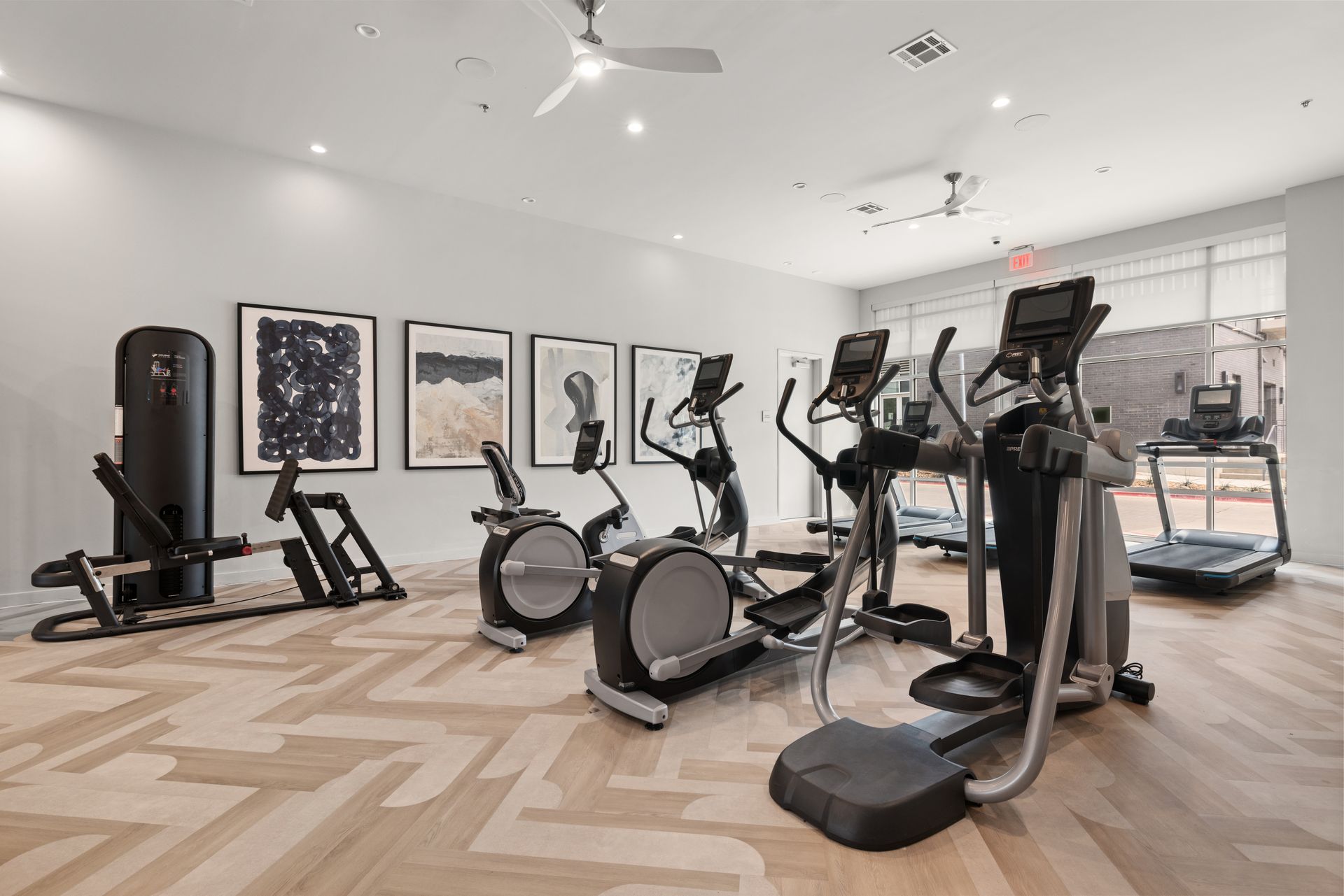 A modern, well-lit fitness center with gray carpeted floor, featuring various exercise machines including stationary bikes, elliptical trainers, and treadmills. The room has white walls adorned with large framed abstract artworks and is equipped with recessed ceiling lights. Large windows with blinds provide natural light and a view to the exterior at 44 South in Austin, TX.