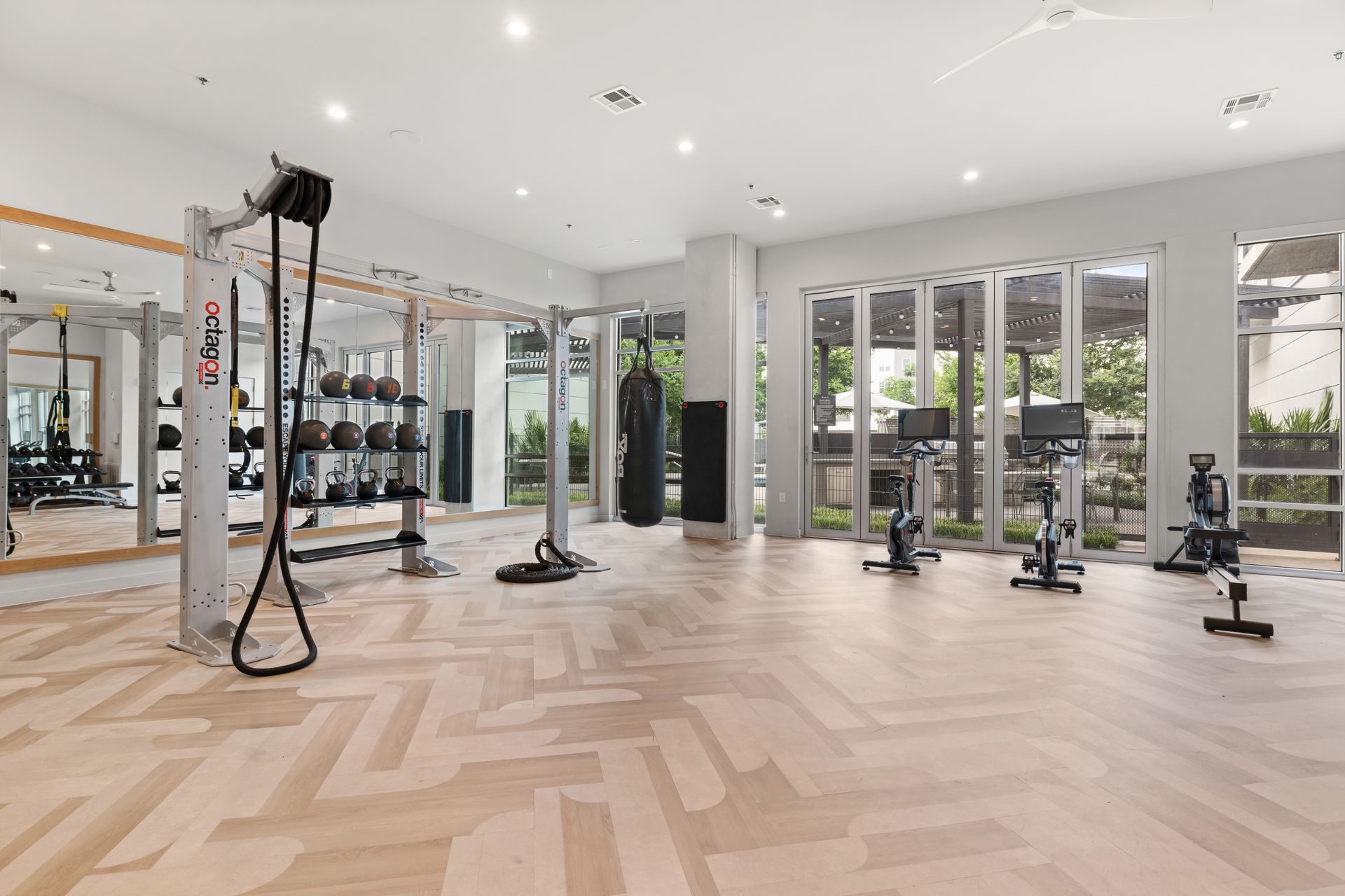 A well-lit, modern gym with large windows offering natural light. The gym features various fitness equipment, including a functional training rig with resistance bands and exercise balls, a punching bag, and several stationary bikes. The floor is tiled with a checkerboard pattern and the space is clean and unoccupied, ready for a workout session at 44 South in Austin, TX.