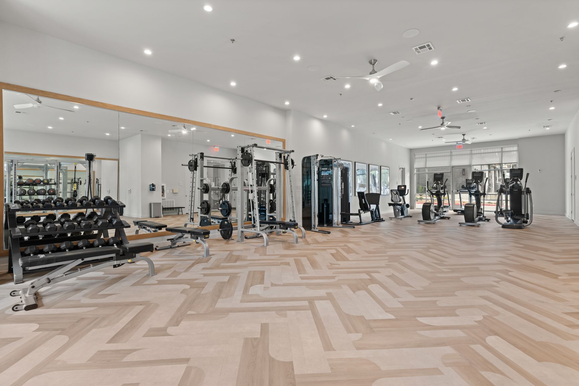 Spacious and modern gym interior with organized workout stations, including a rack of dumbbells, adjustable benches, and various exercise machines. The room has a large mirror along one wall, bright lighting, and a checkerboard pattern floor at 44 South in Austin, TX.