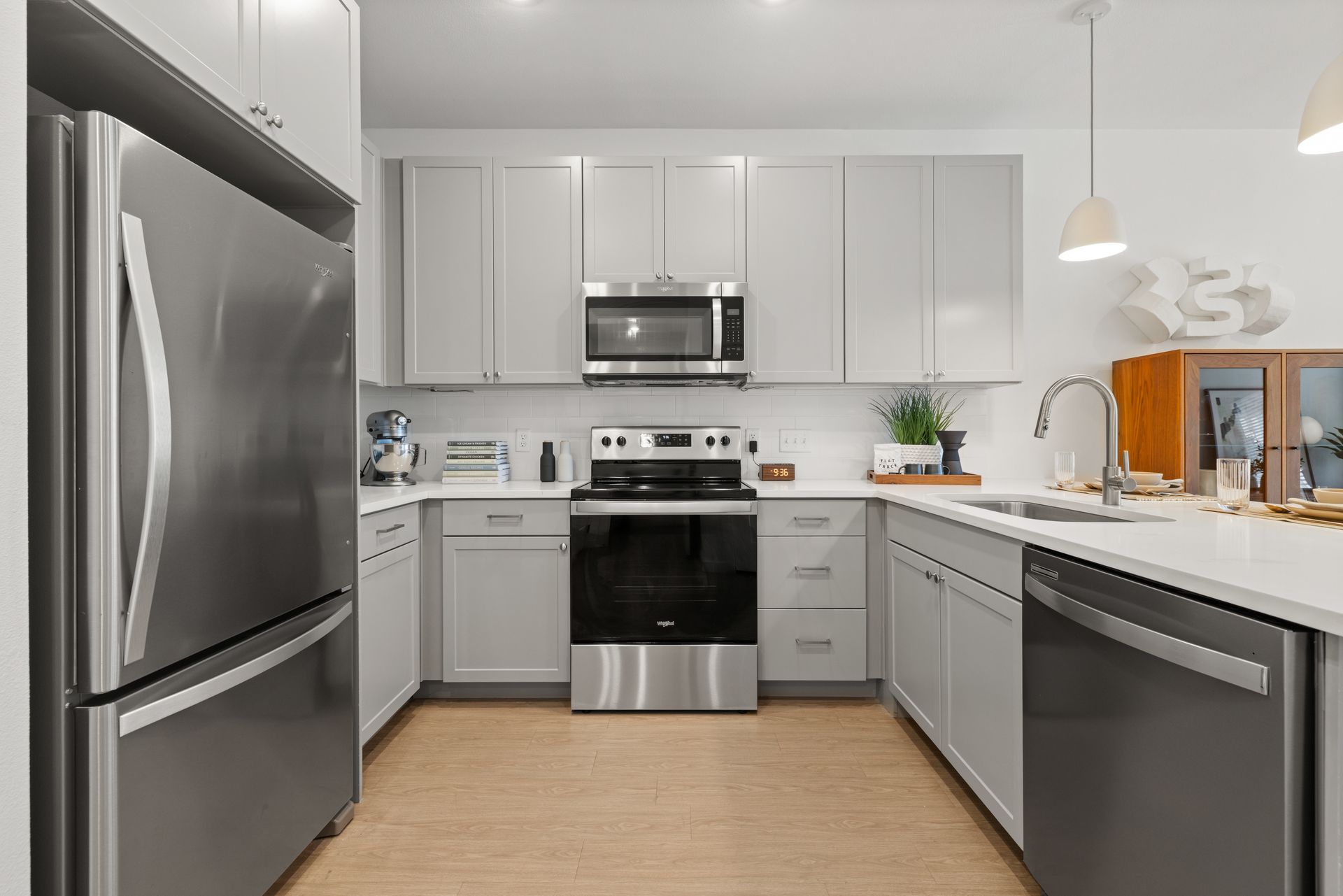 Modern kitchen interior with stainless steel appliances, light grey cabinets, and a teal accent wall. There is a standing refrigerator, a microwave over a range, and a dishwasher. The kitchen includes a light wood floor, white countertops, and a hanging white lamp. Decorative touches include a stand mixer, plants, and neatly arranged books and kitchenware at 44 South in Austin, TX.