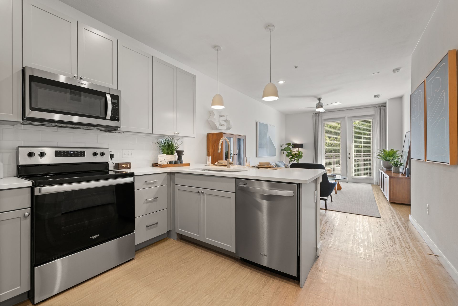 Modern kitchen with white cabinetry, stainless steel appliances, and an island extending into a cozy living area with a sofa, chairs, and a television. The space is brightly lit with natural light from a balcony door and features a combination of soft teal and white walls, accented with framed wall art at 44 South in Austin, TX.