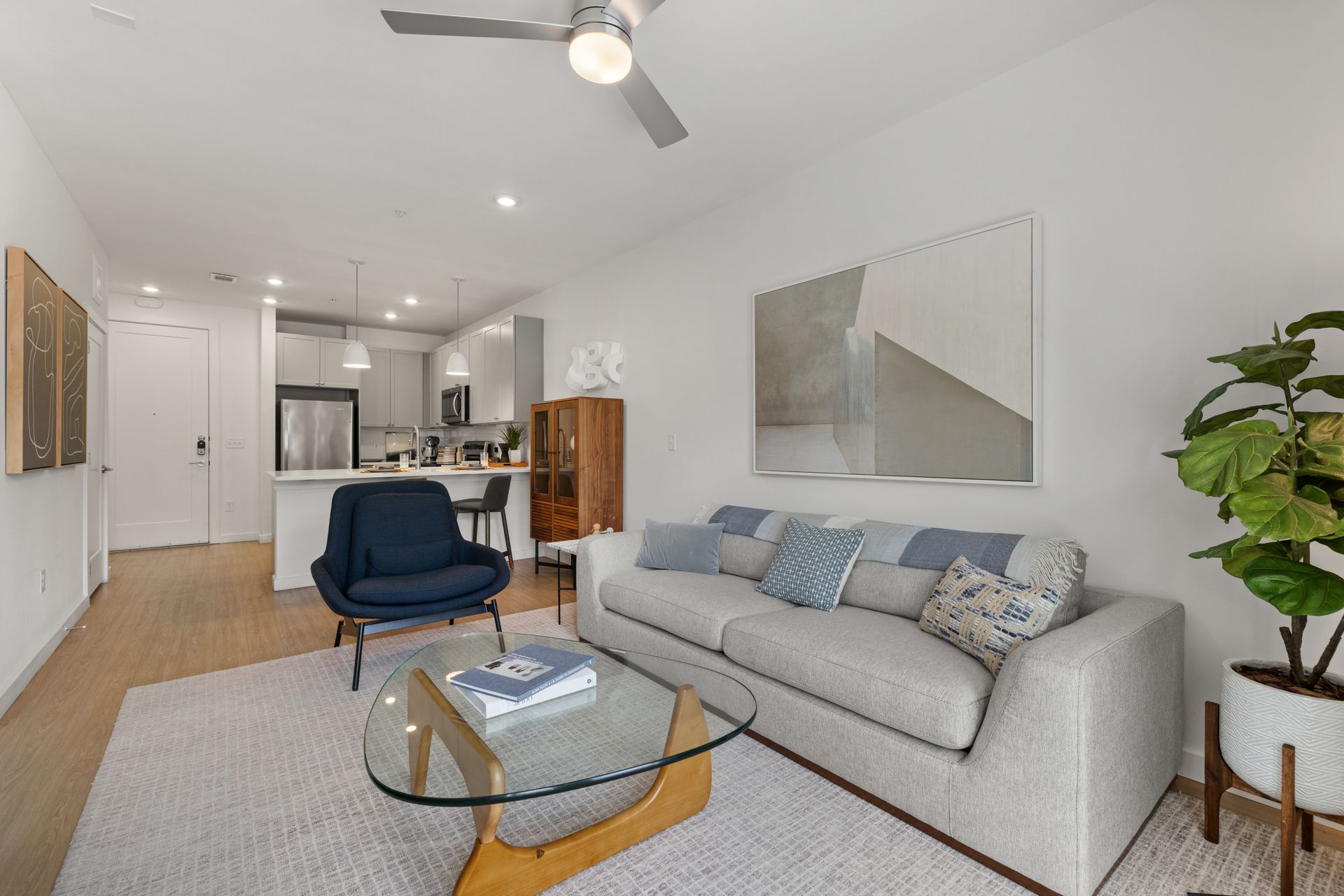 Modern living room interior with an open floor plan featuring a lounging area with a beige couch, blue accent chair, and a glass coffee table on a textured rug. A blue accent wall with a large abstract painting complements the white walls. The connected kitchen appears in the background with white cabinets and stainless steel appliances. No people are visible at 44 South in Austin, TX.