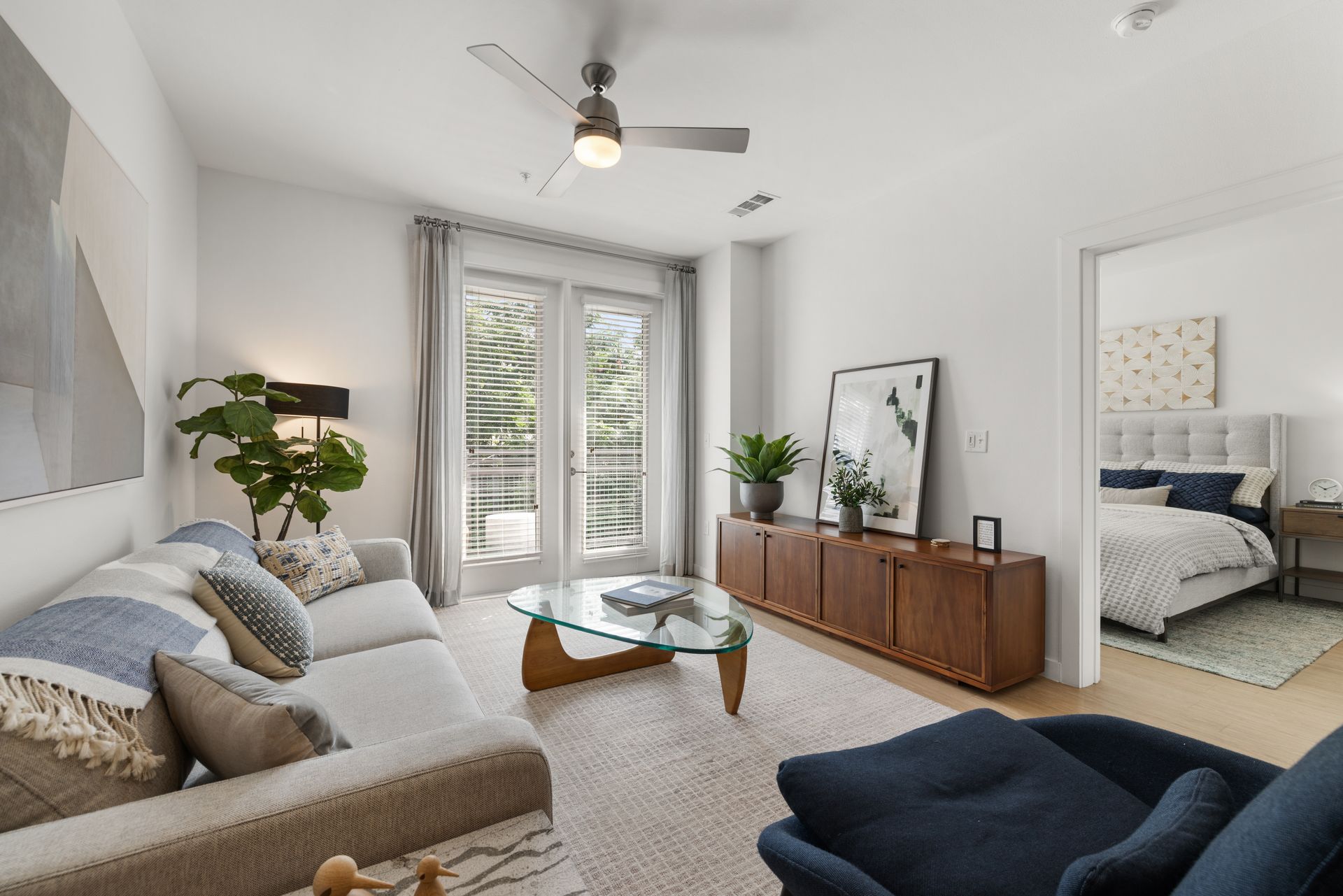 Modern living room interior in neutral tones with a beige sofa, blue accent chairs, and a mid-century style wooden sideboard. A large abstract painting hangs on the accent wall, and through the glass door, a balcony with greenery is visible. The room opens to a cozy bedroom with a tufted headboard at 44 South in Austin, TX.