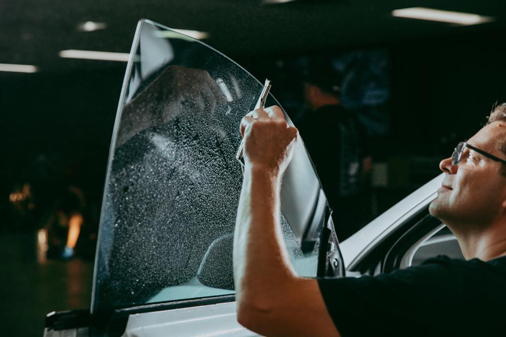 Person applying tint film to a car window in a dark garage, holding a squeegee near a cracked-looking sheet