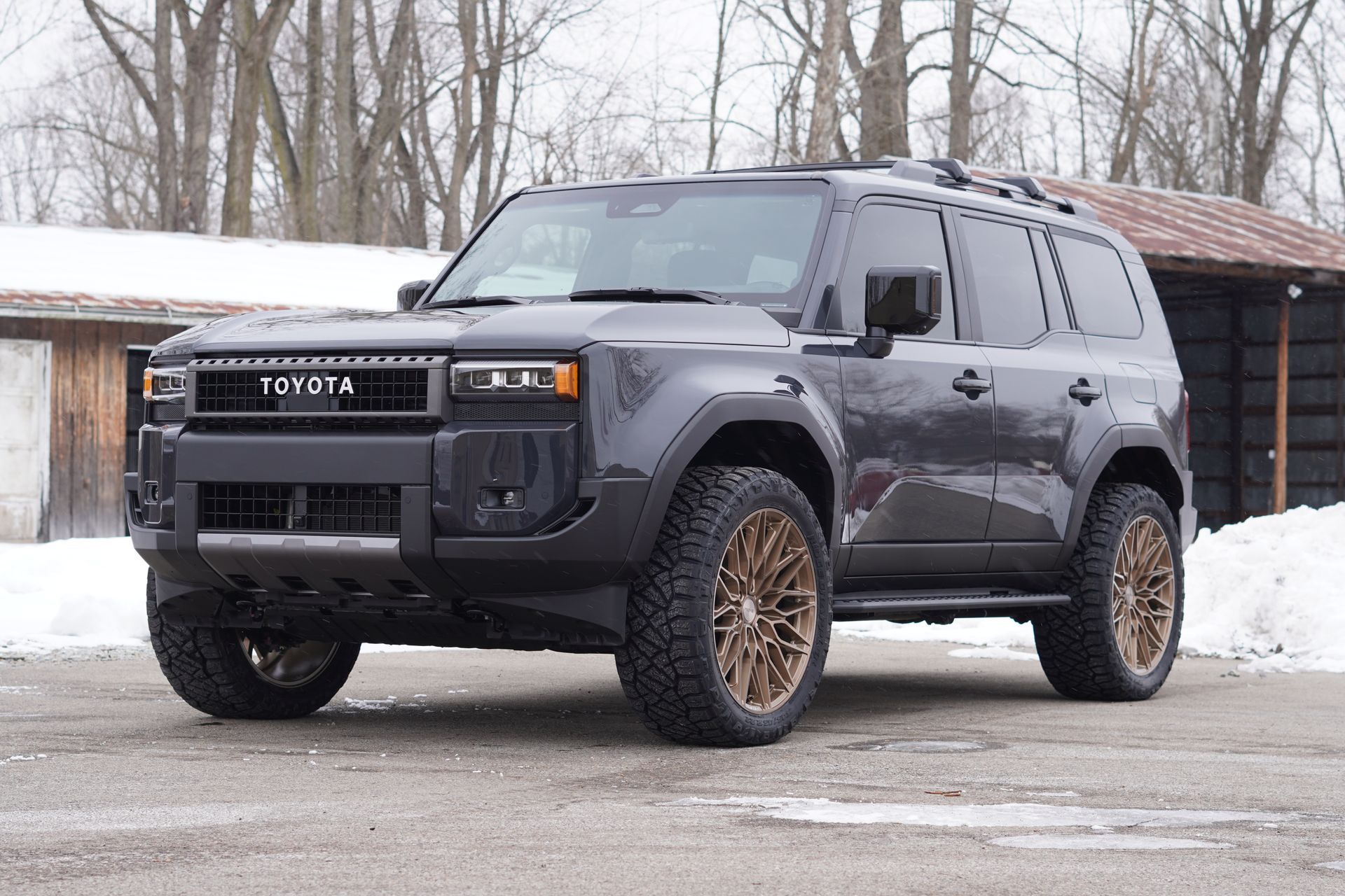 Dark gray SUV with bronze wheels parked in a snowy area, near a building.