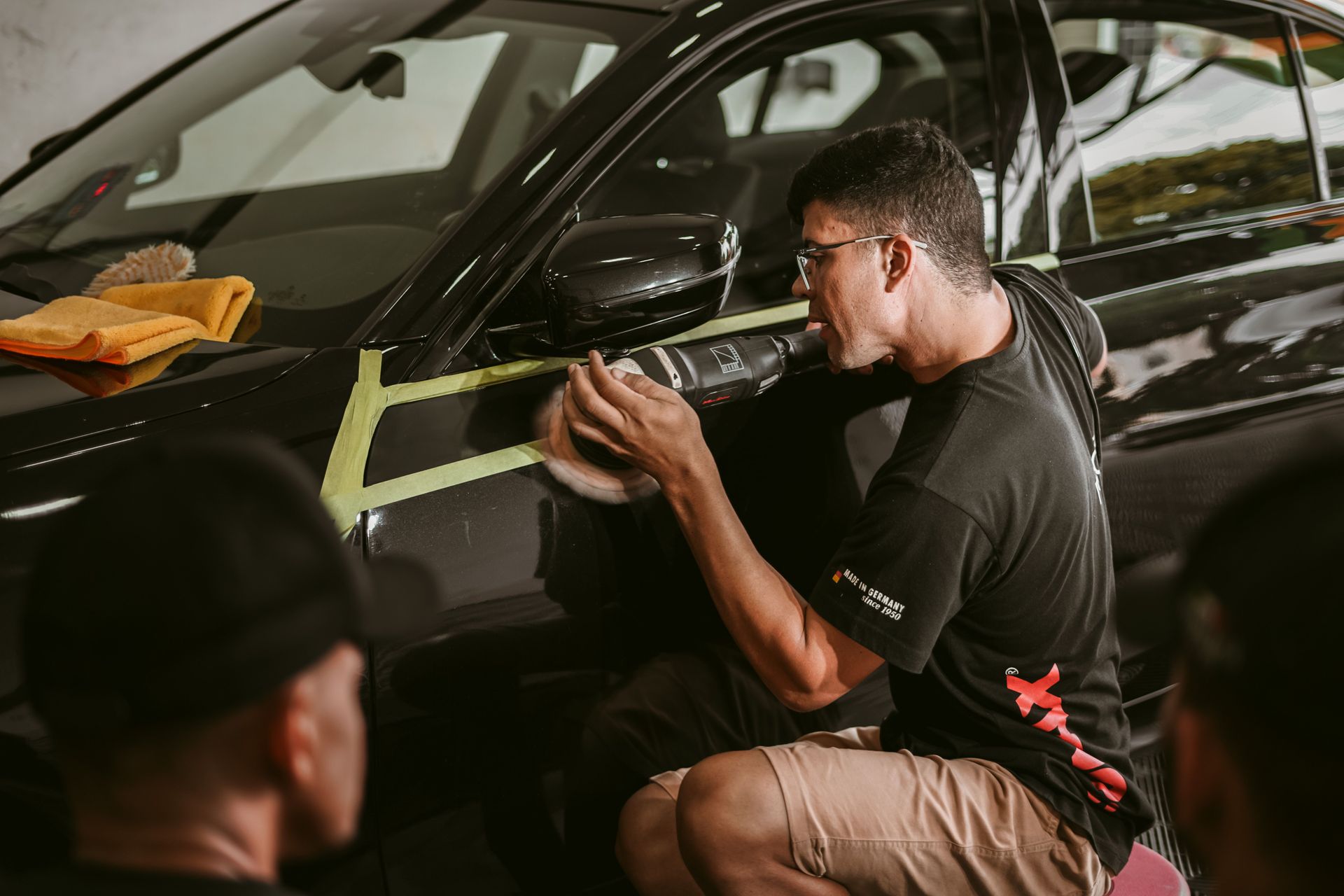 Two workers polishing a black car with yellow tape marking the repair area in a shop