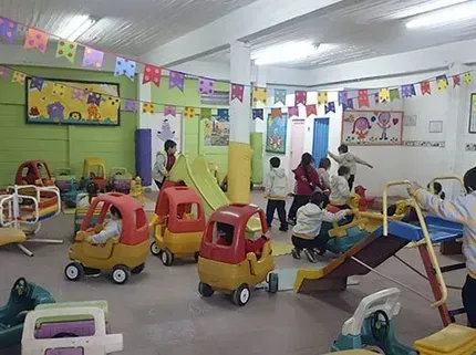Sala de juegos interior de guardería donde los niños montan en coches de juguete y juegan en toboganes y colchonetas.