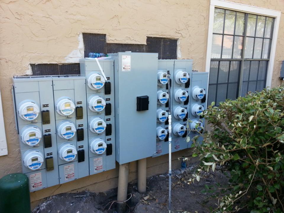 Outdoor utility meters and electrical boxes mounted on a beige wall beside a window and shrub.