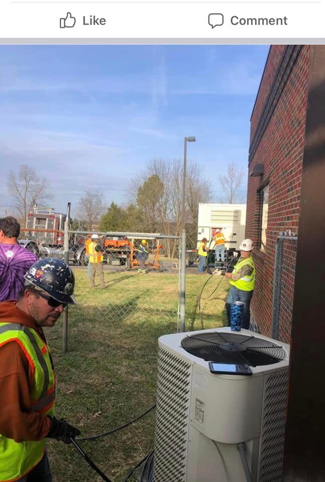 Workers in safety vests and hard hats near a brick building and outdoor equipment under a blue sky
