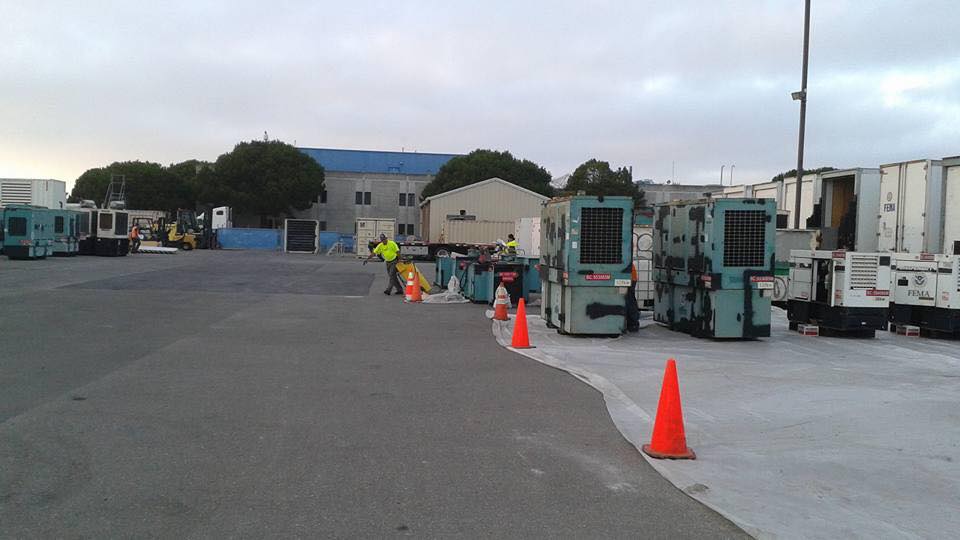 Industrial yard with parked machinery, orange cones, and workers in safety vests near a warehouse