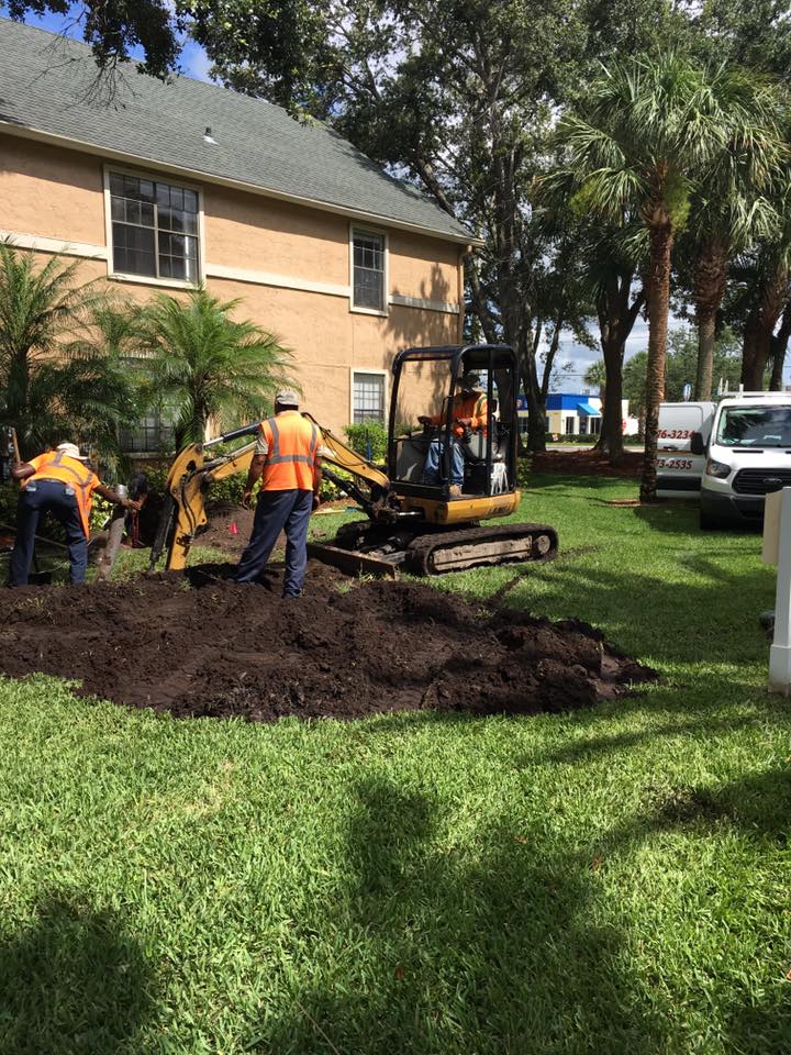 Workers in orange vests use a mini excavator to move mulch in a grassy yard by an apartment building.