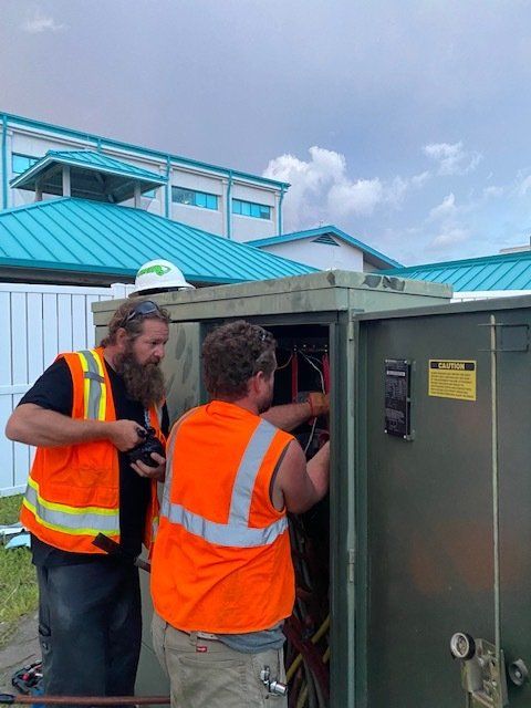 Two utility workers in orange safety vests inspect an outdoor electrical cabinet on a rooftop.