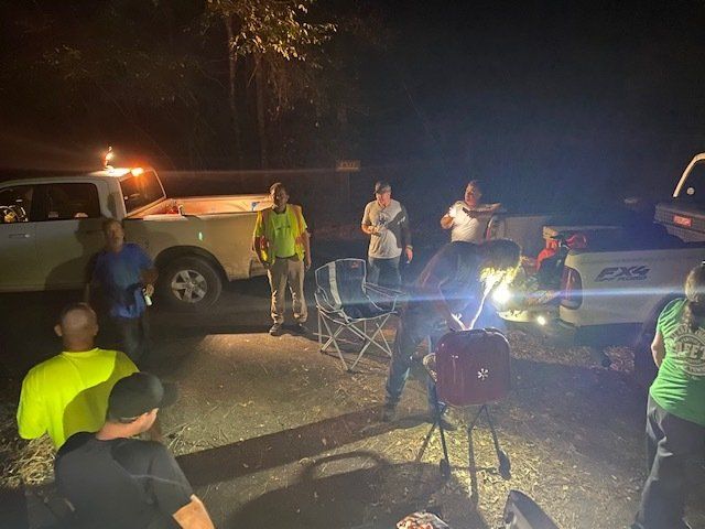 Night roadside scene with several people gathered around two pickup trucks and bright work lights on a dim street