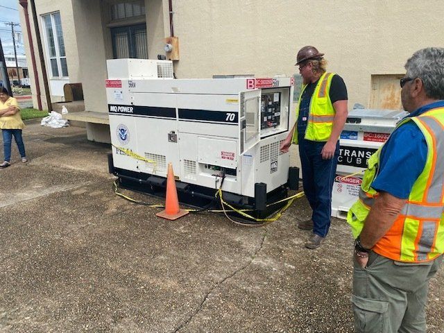 Workers inspect a white utility trailer beside a building, with orange cones and safety vests.