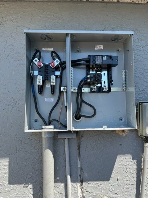 Open outdoor electrical control box with wiring, breakers, and conduit mounted on a wall.