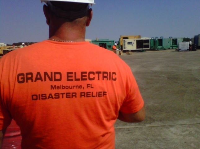 Worker in orange shirt labeled “Grand Electric Disaster Relief” at a disaster site with parked trucks.