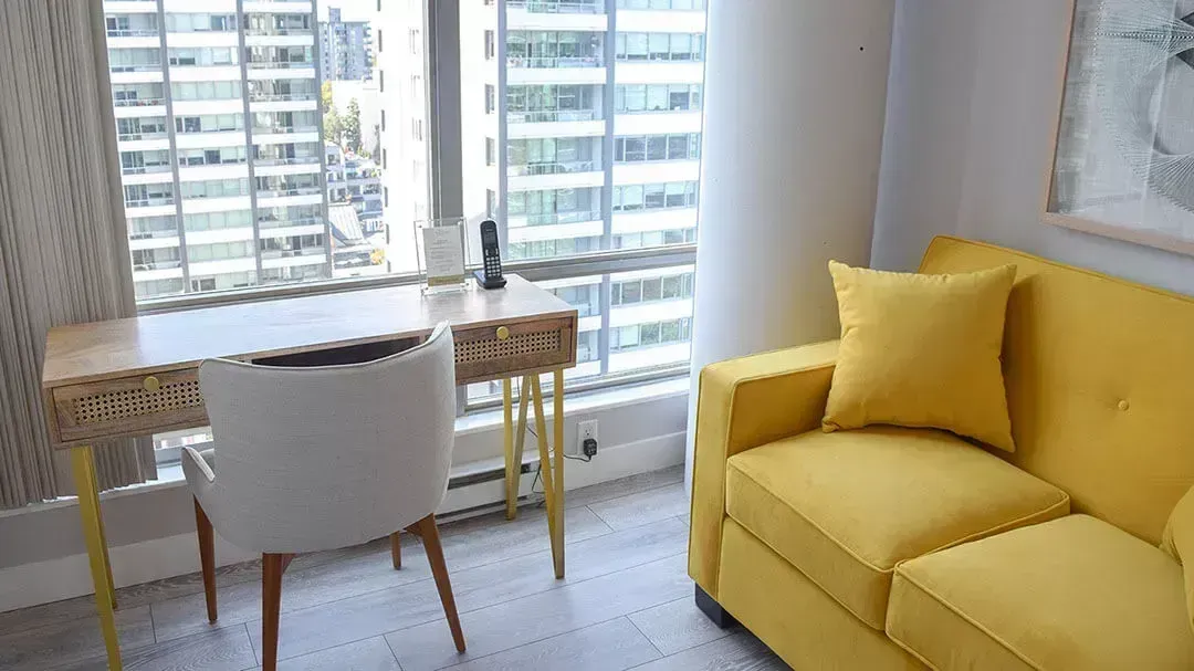 Yellow sofa pillow on top of a yellow sofa next to a wooden table, wooden chair, and a window