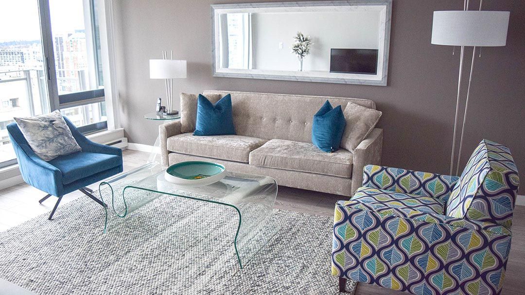 Two blue sofa pillows on top of a beige sofa next to a glass coffee table, a mirror, and a sofa chair with blue and yellow wavy design
