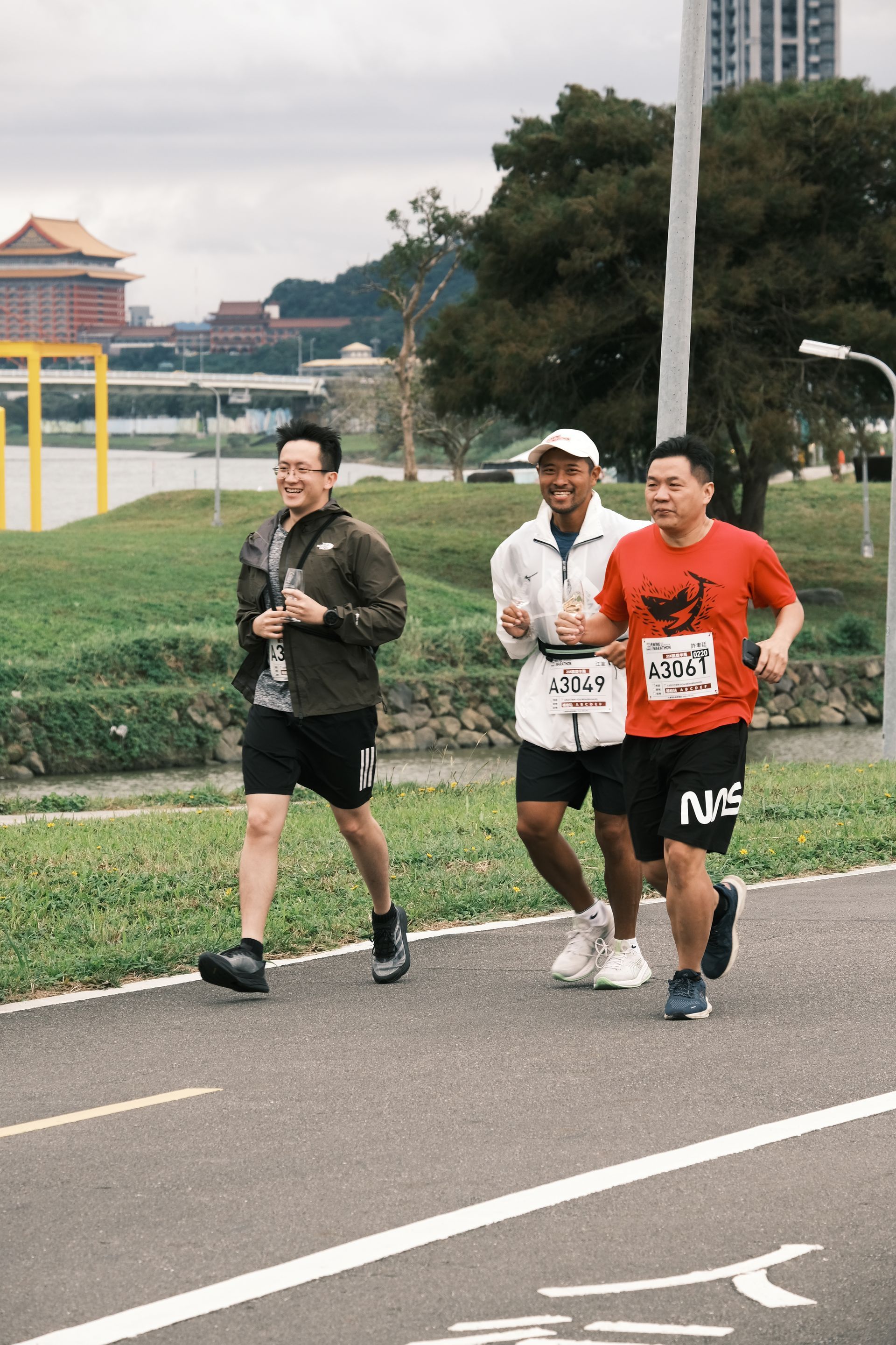 三名跑者沿著道路慢跑，旁邊是綠草如茵的公園和鐵絲網圍欄。
