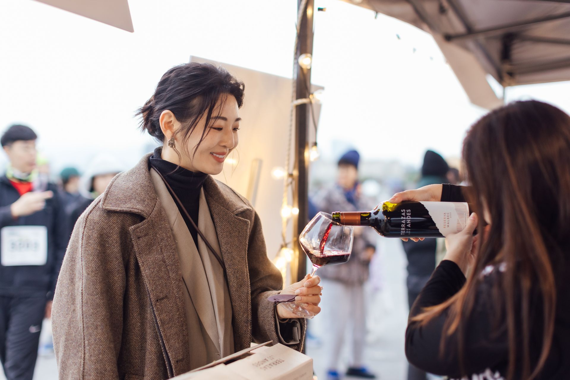 一名穿著棕色外套的女子拿著酒杯，另一人在戶外活動中倒酒給她。