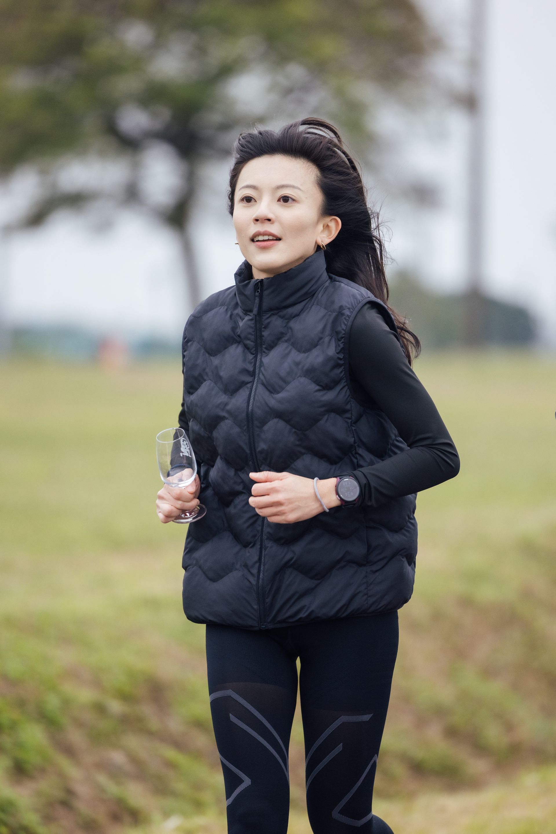 一名穿著黑色背心和緊身褲的女子，手拿水壺在草地小徑上慢跑