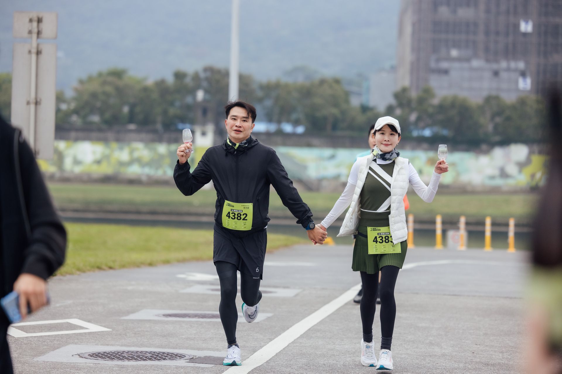 兩名穿著黑色運動服的跑者在公園道路上跑步時揮手，身上的號碼布清晰可見。