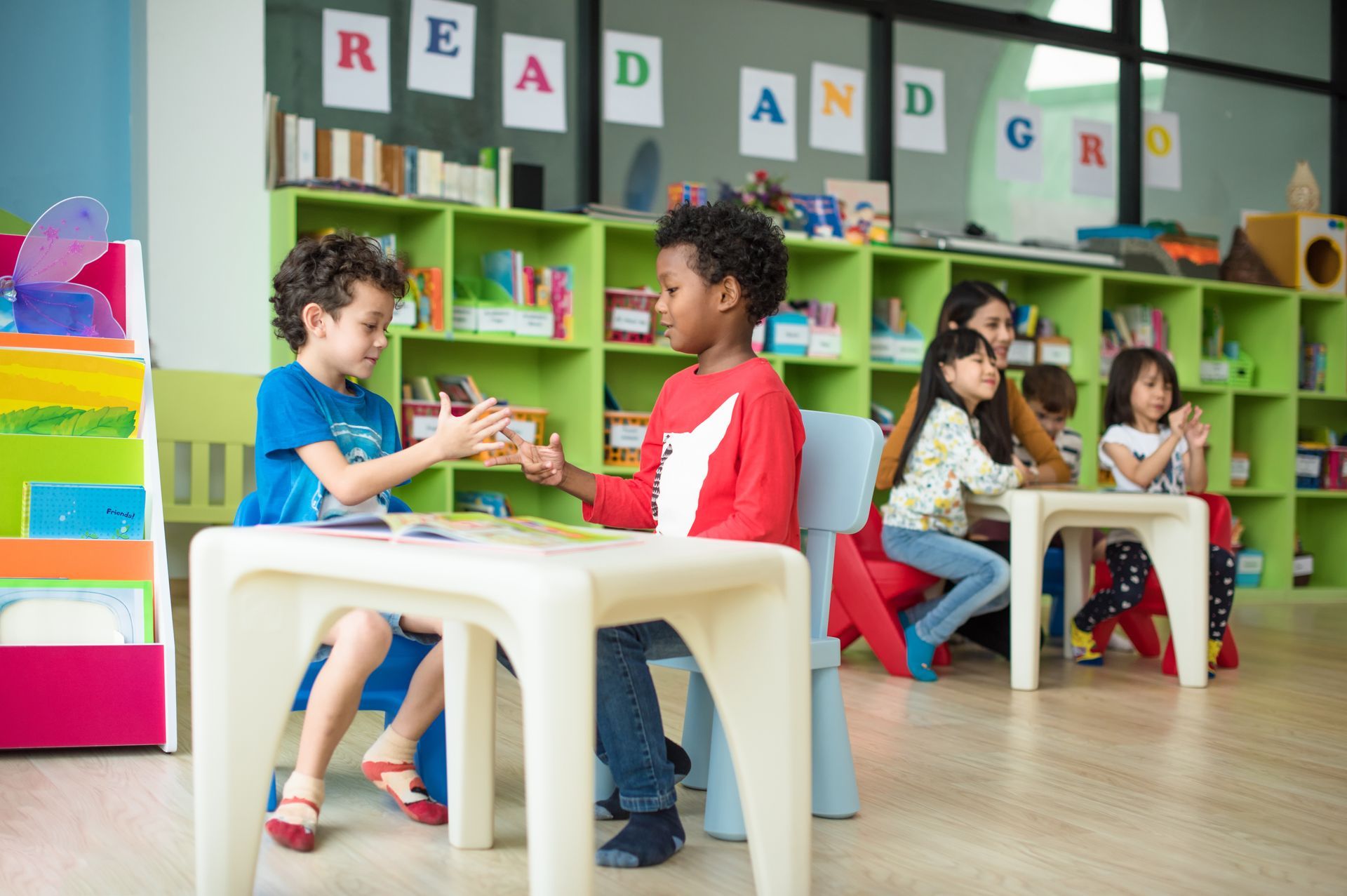 Two young preschool boys sit at a small white table interacting while classmates read books behind Two young preschool boys sit at a small white table interacting while classmates read books behind