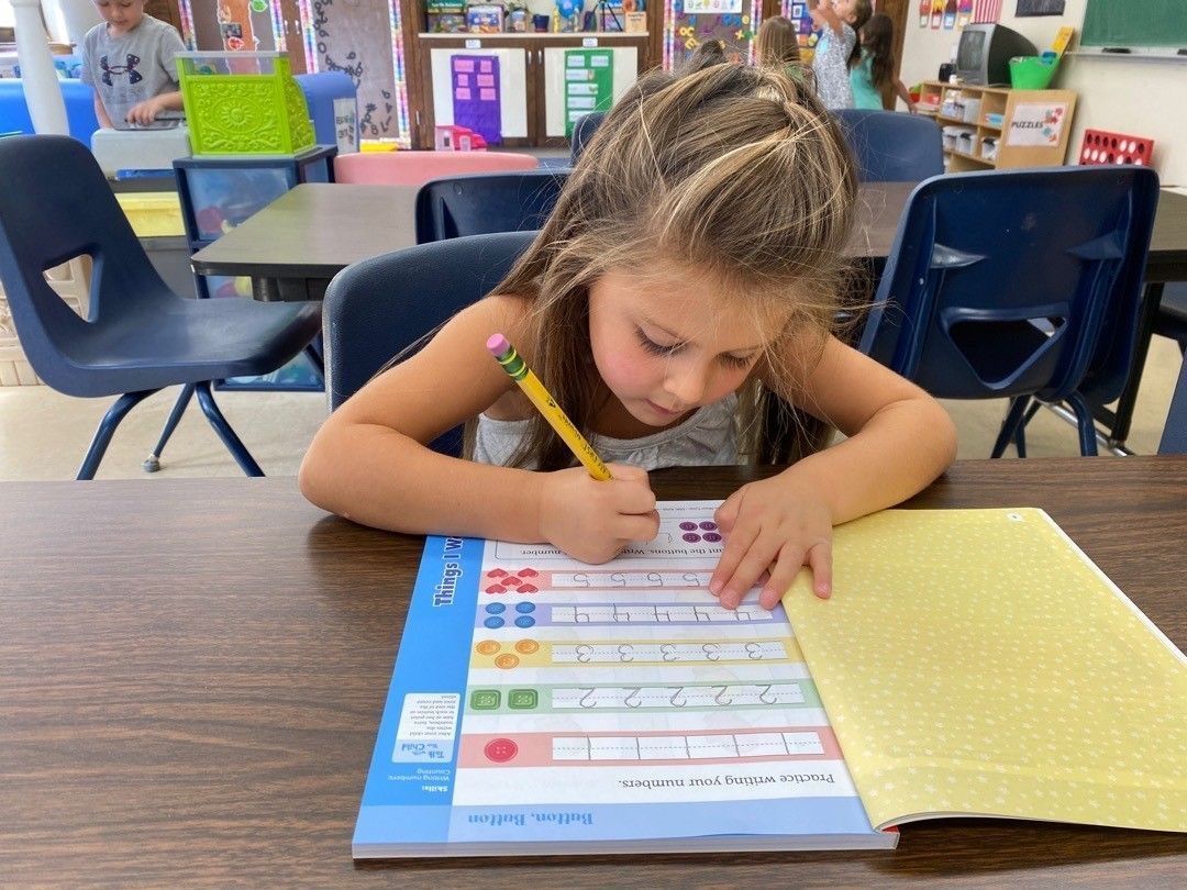 Girl writing at a desk in a classroom. She uses a pencil, focused, with a worksheet.