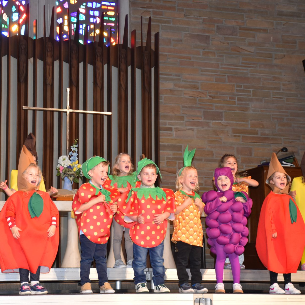 Children in colorful fruit costumes standing on a stage in front of a church organ
