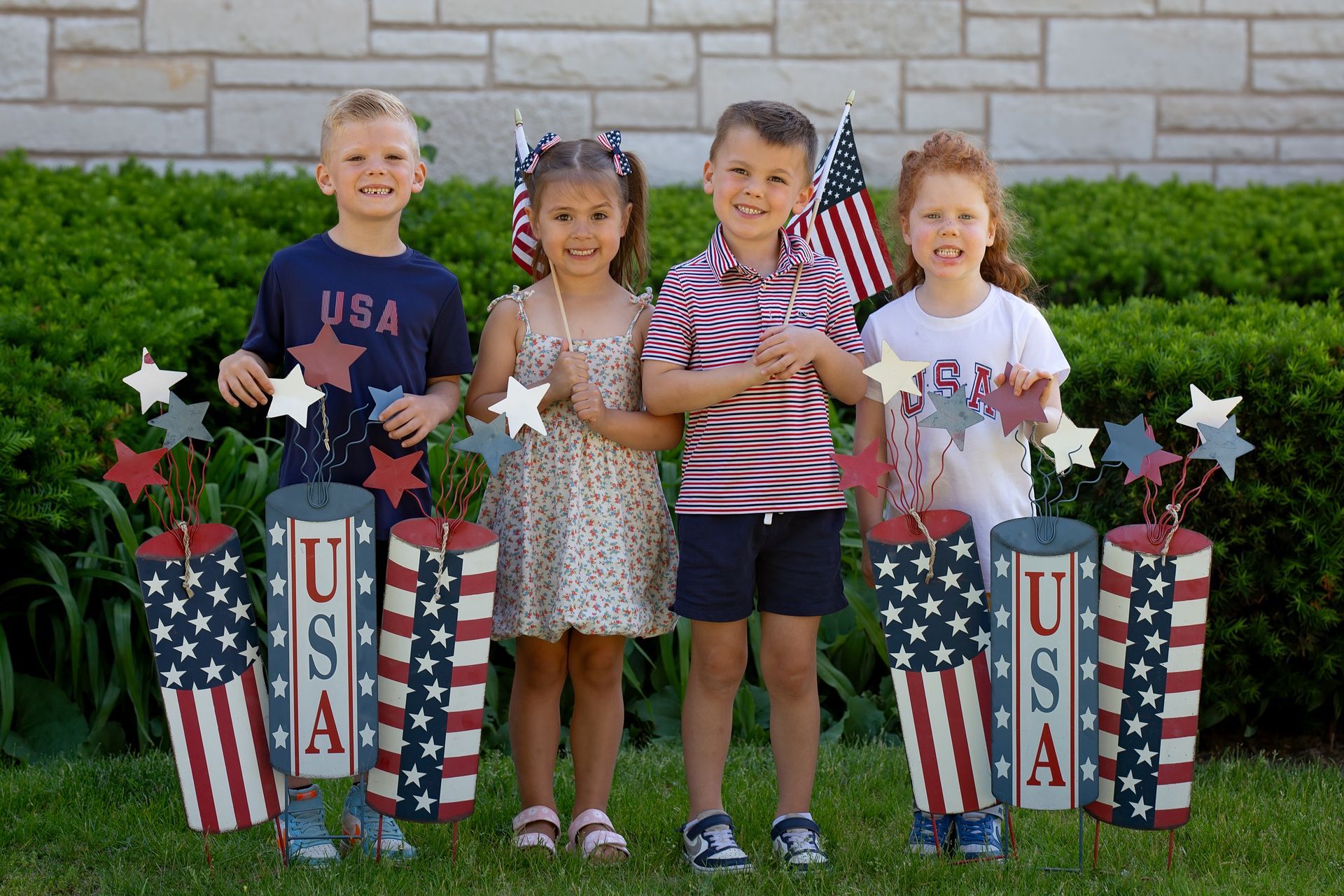 Four children standing on grass beside patriotic USA decorations and stars, smiling at the camera.