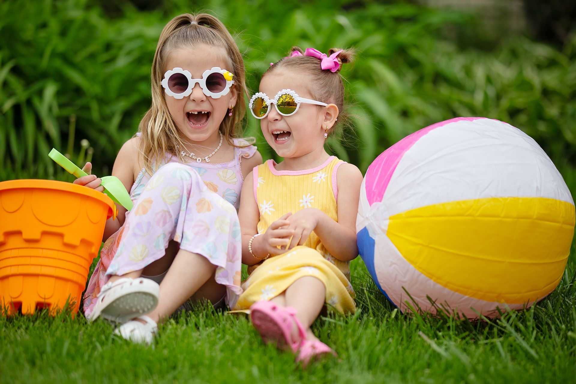 Two children in sunglasses sitting on grass with a beach ball and orange bucket outdoors