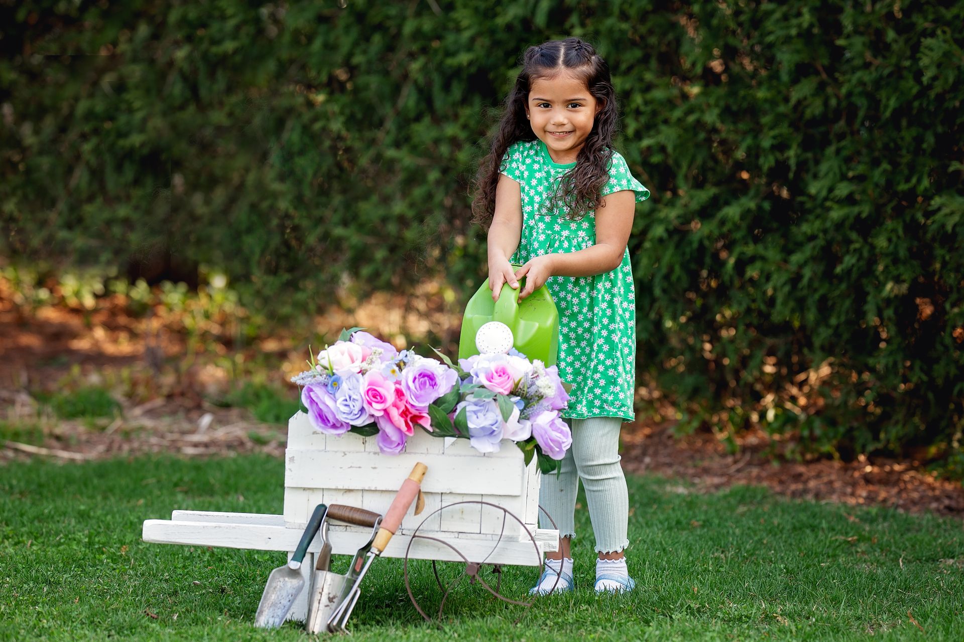 Young girl in a green dress stands beside a white cart with purple flowers on a grassy lawn.
