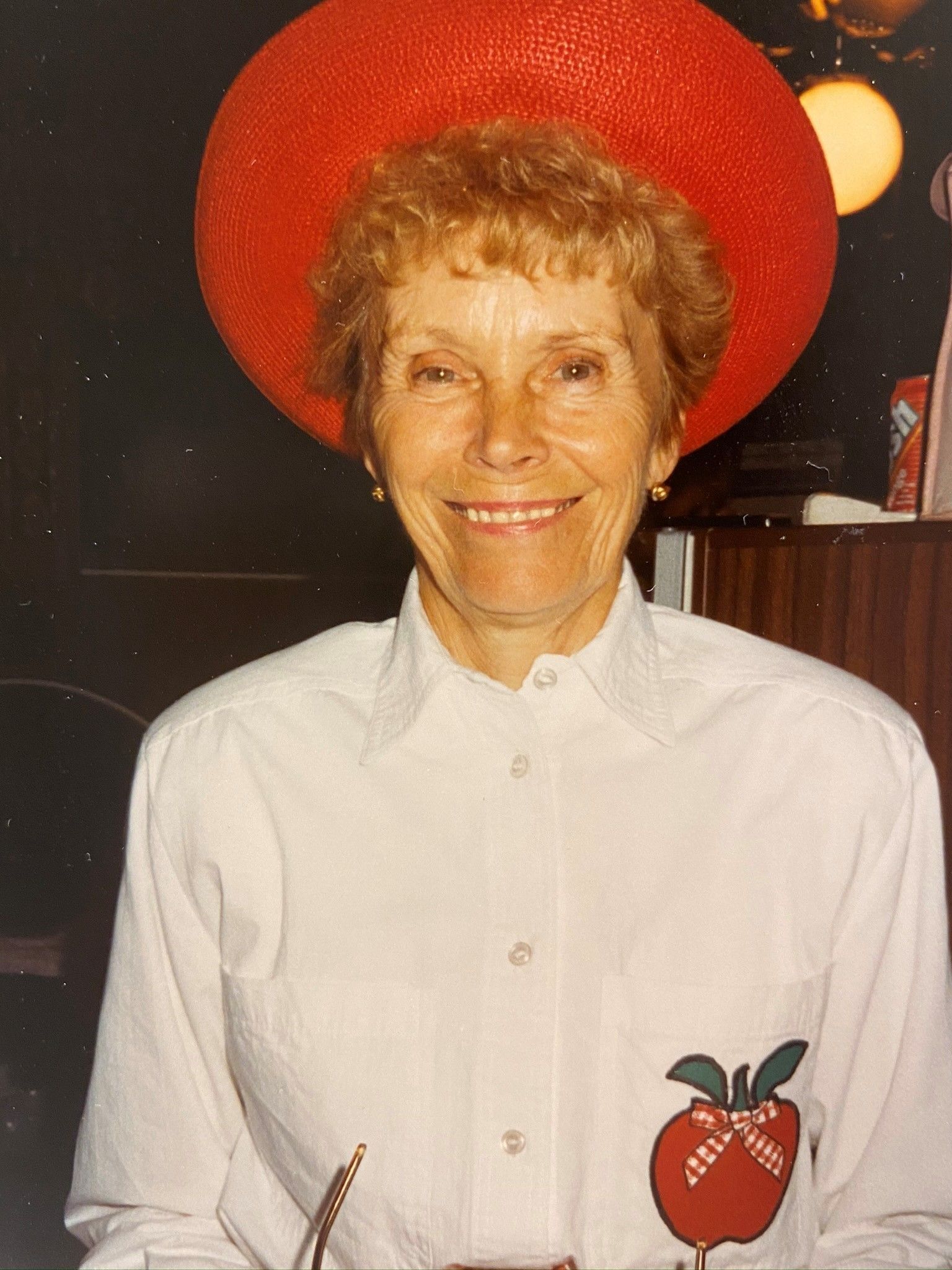 Woman wearing a large red hat and white shirt with an apple design, smiling.