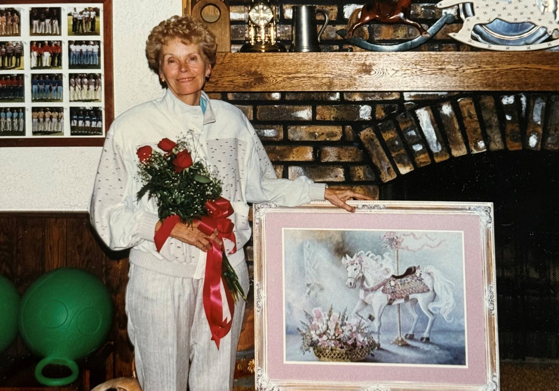 Woman holding roses and artwork of a white horse, standing by a brick fireplace.