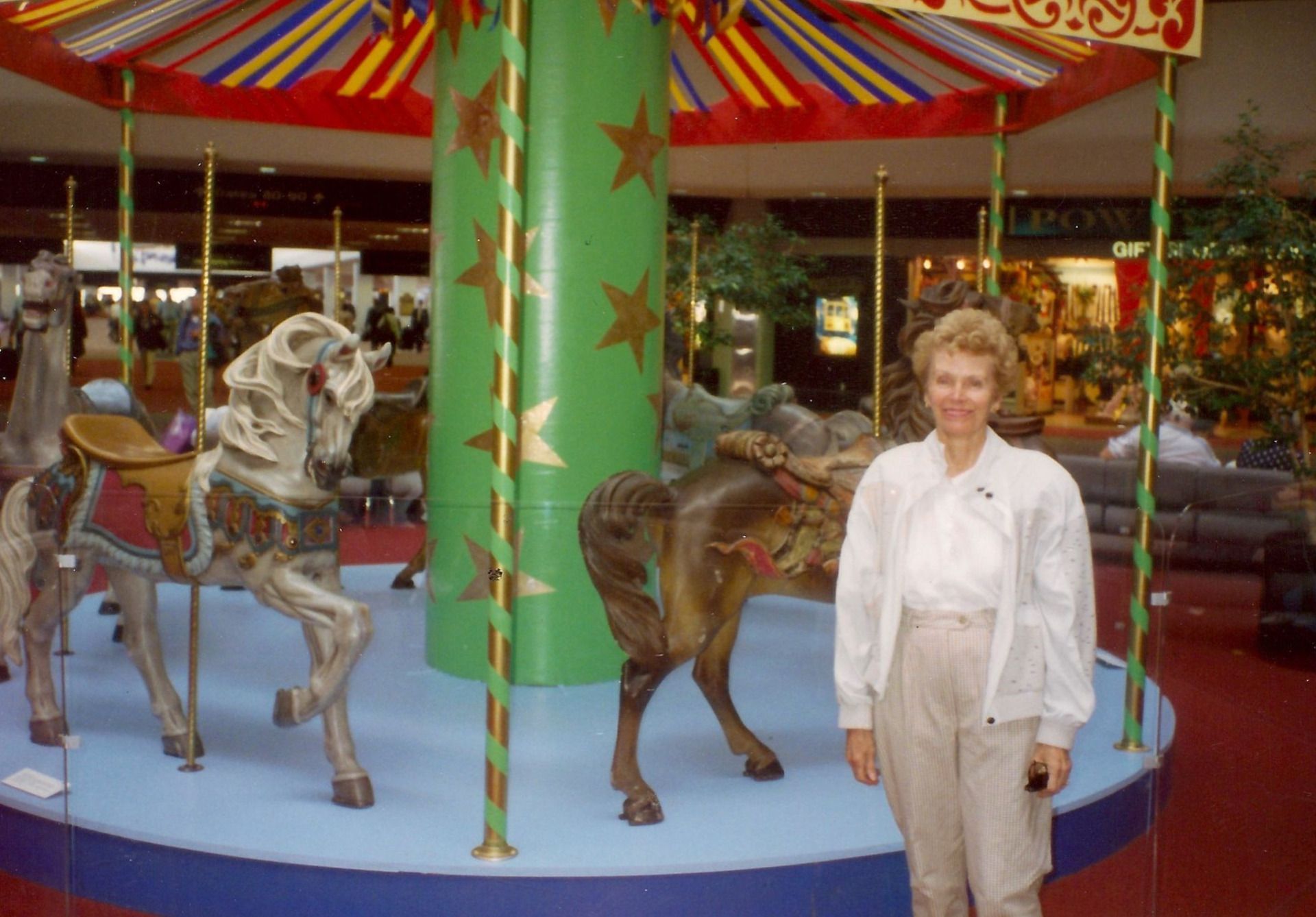 Woman stands next to a carousel in a mall. Carousel has horses and a green column.