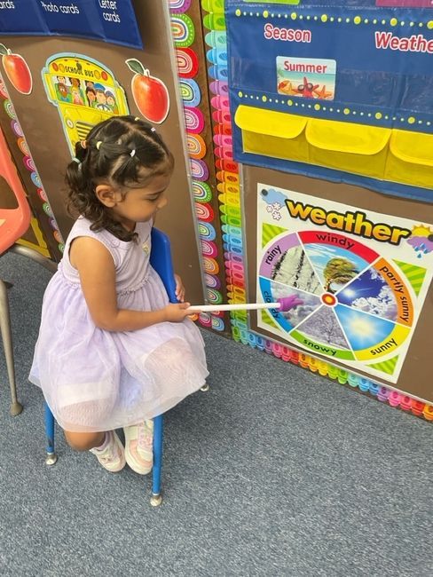 Child in a white dress points at a colorful weather wheel in a classroom.