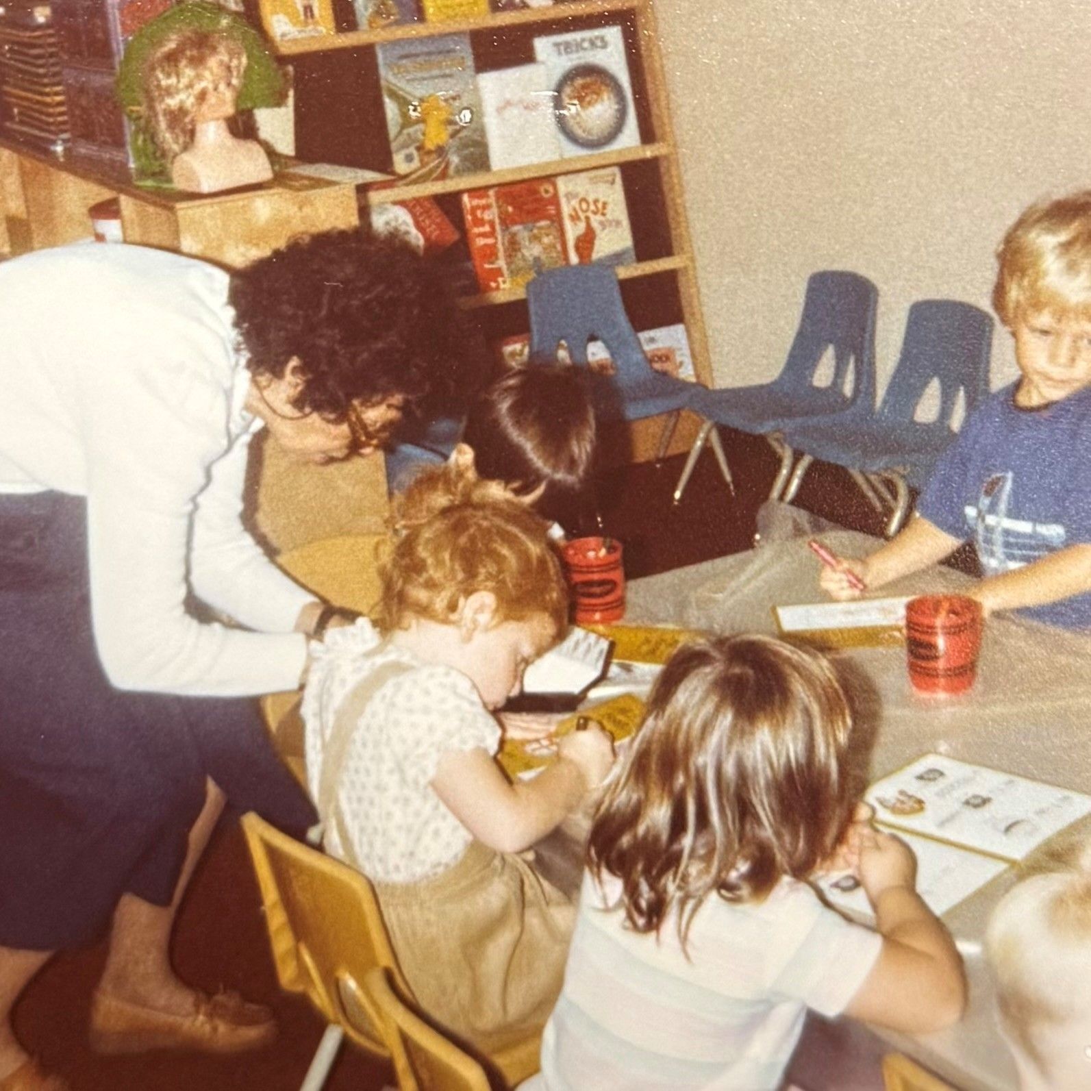 Teacher helping young children at a table, drawing. Classroom with bookshelves in the background.
