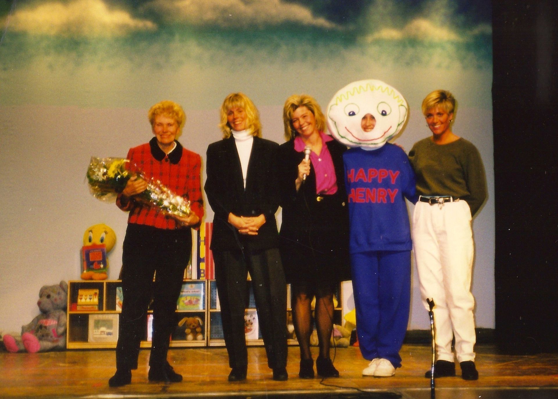 Four women and a mascot on stage; one holds flowers. Blue backdrop with clouds.