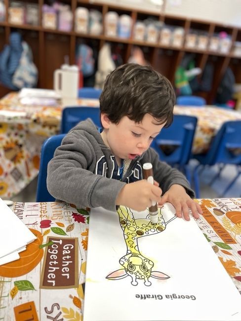 Child coloring at a table with a dinosaur worksheet in a classroom, focused and leaning forward.