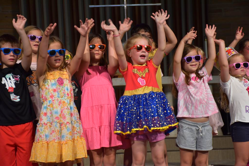Children on stage with hands raised, wearing colorful clothes and sunglasses