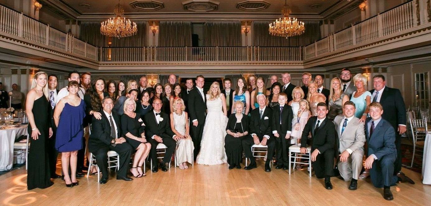 Large group photo at a wedding reception. People in formal attire pose in a ballroom.