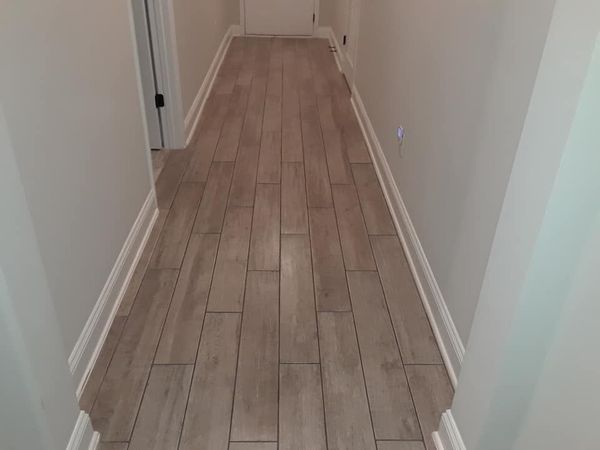A narrow hallway with light-toned, wood-grain floor tiles, white baseboards, and neutral-colored walls.