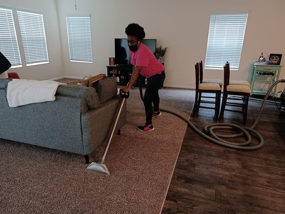 A person in a pink shirt cleans a carpeted living room with a vacuum wand near a grey sofa.