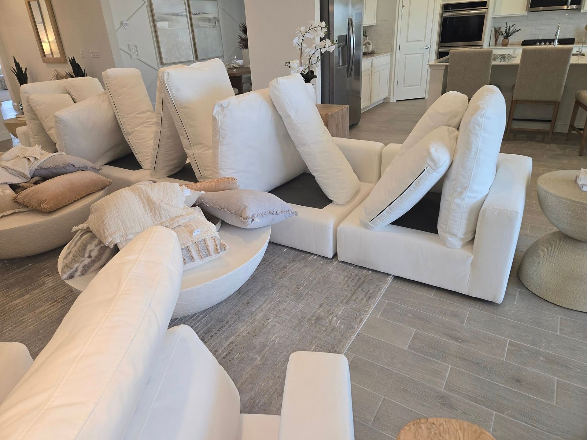 A bright, modern living room featuring a modular white sofa with several pillows, set on a light rug near an open kitchen.