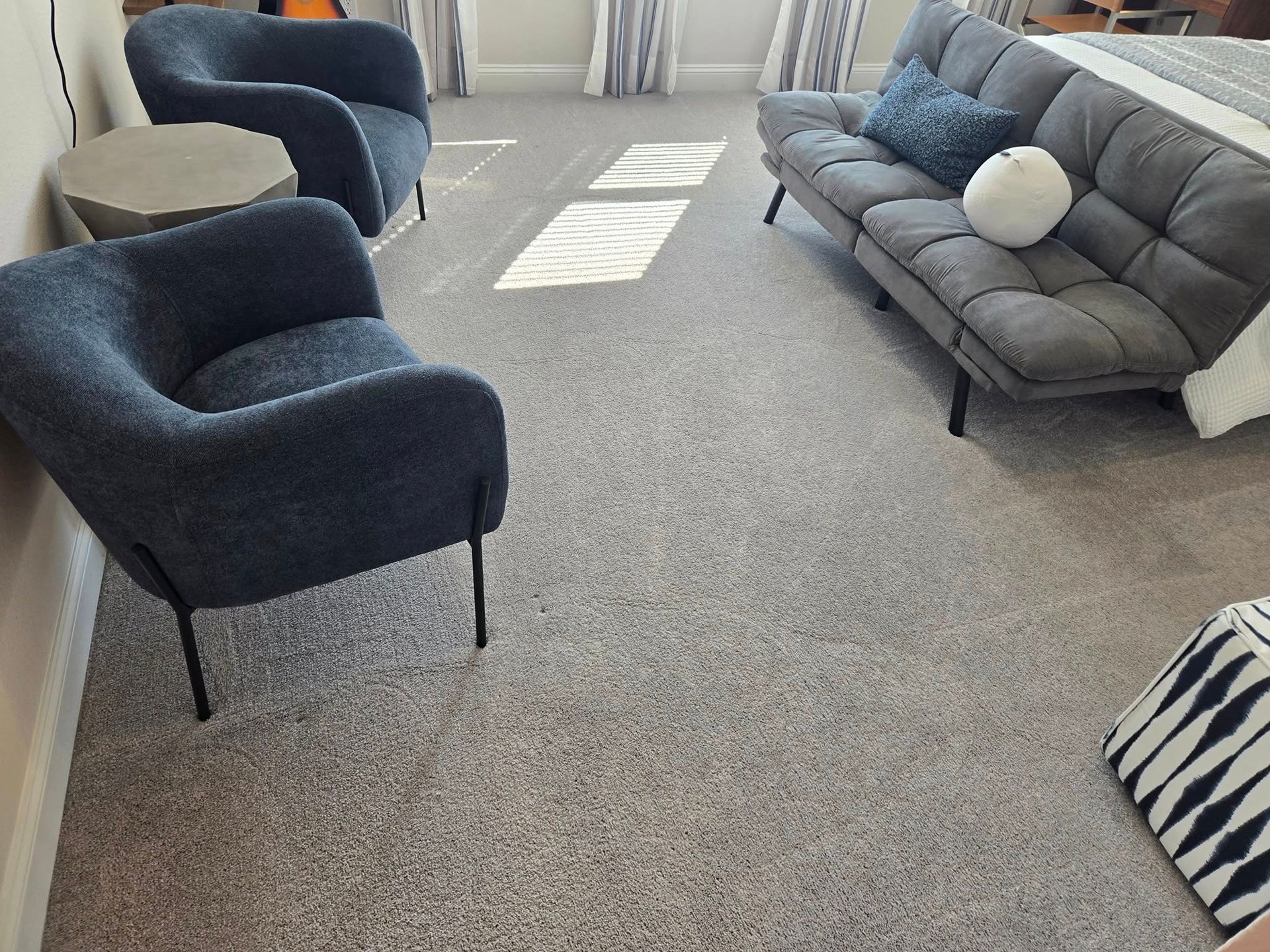 A room with two dark blue textured chairs, a small round table, and a gray sofa with blue and white pillows on carpet.