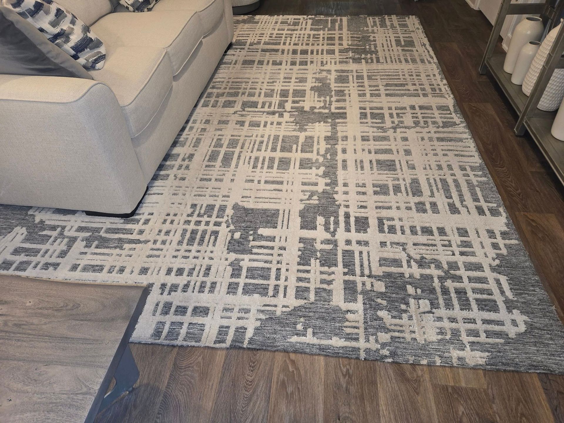 A light gray couch sits on a large, textured rug with a dark gray and cream abstract grid pattern on a wood floor.