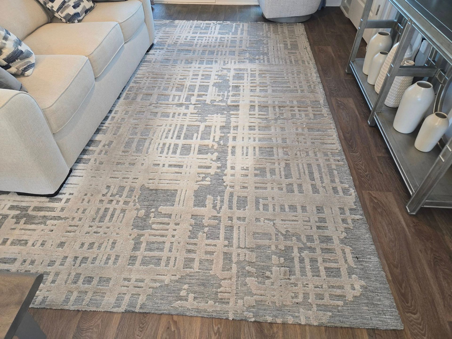 A rectangular area rug with a blue and cream abstract grid pattern sits on a dark wood floor near a sofa and decor.