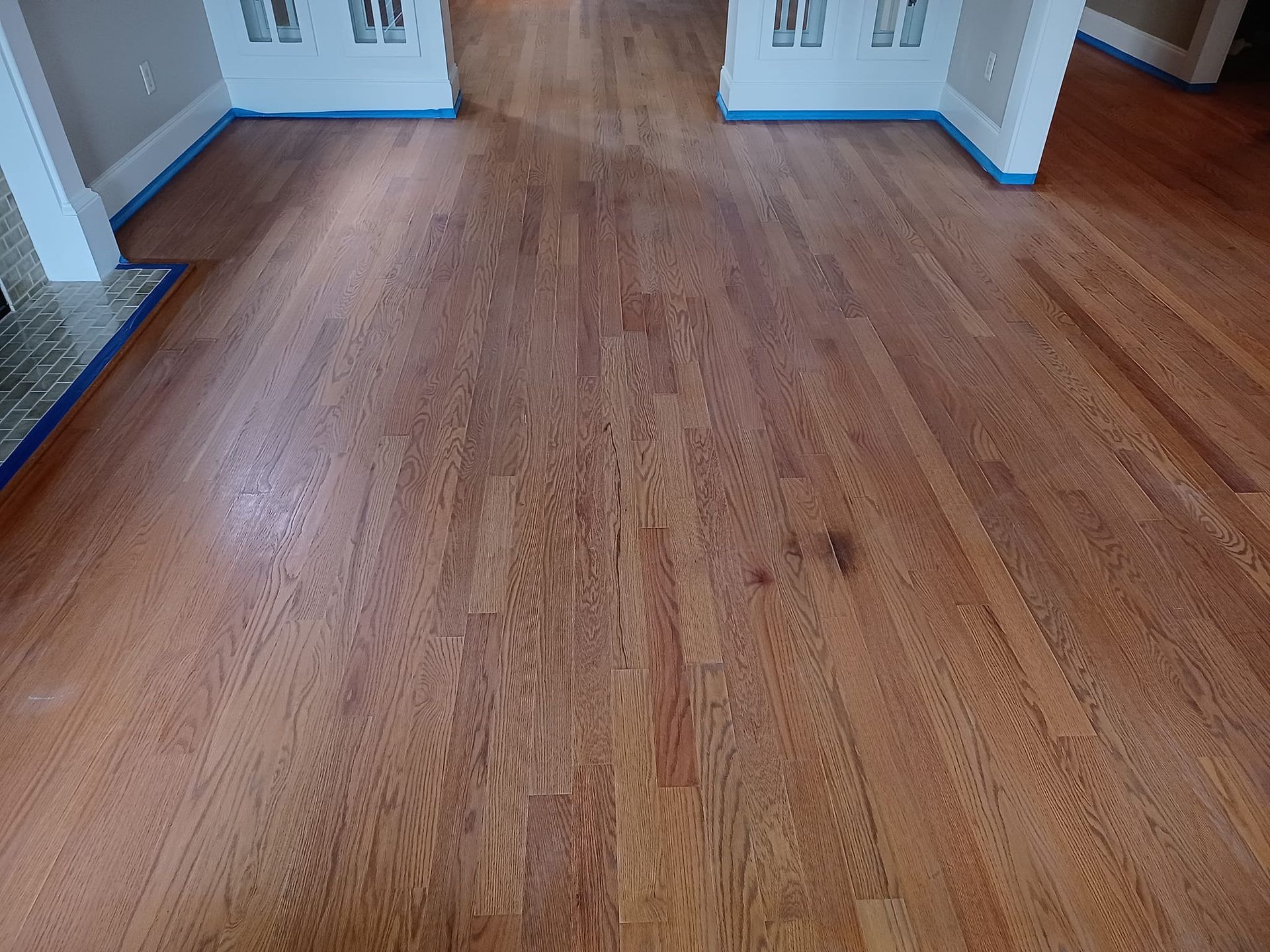 A wide-angle view of light-colored hardwood flooring installed in a room with white baseboards and blue painter's tape.