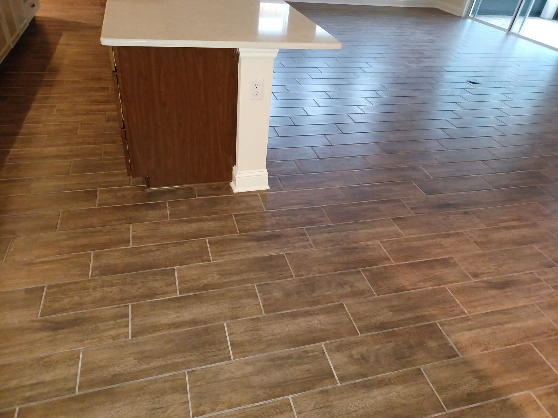A kitchen island with a white countertop stands on a floor covered in brown, wood-look rectangular tiles.